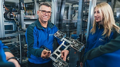 A man and a woman in blue work jackets looking together at a metal component held in his hands