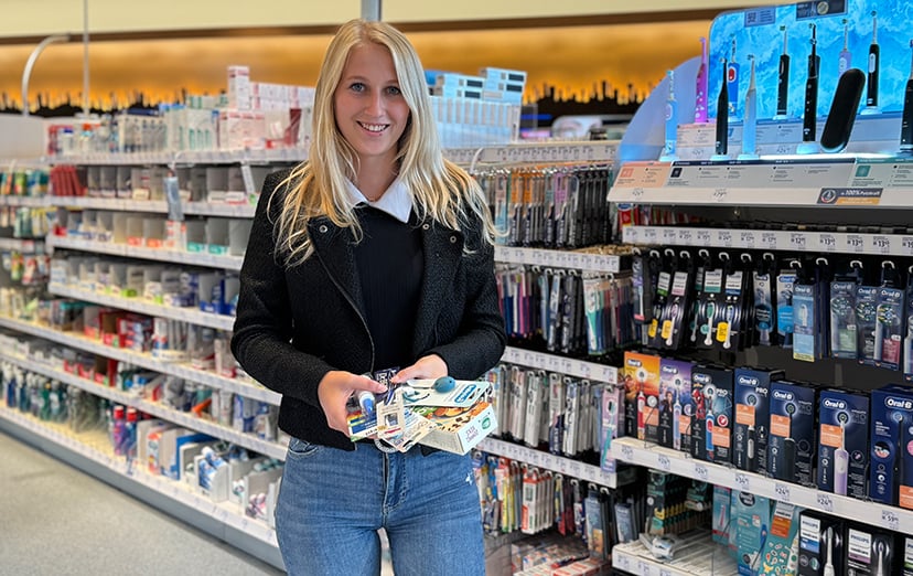 Smiling woman in a shop aisle holding an armful of blister-packed personal care products