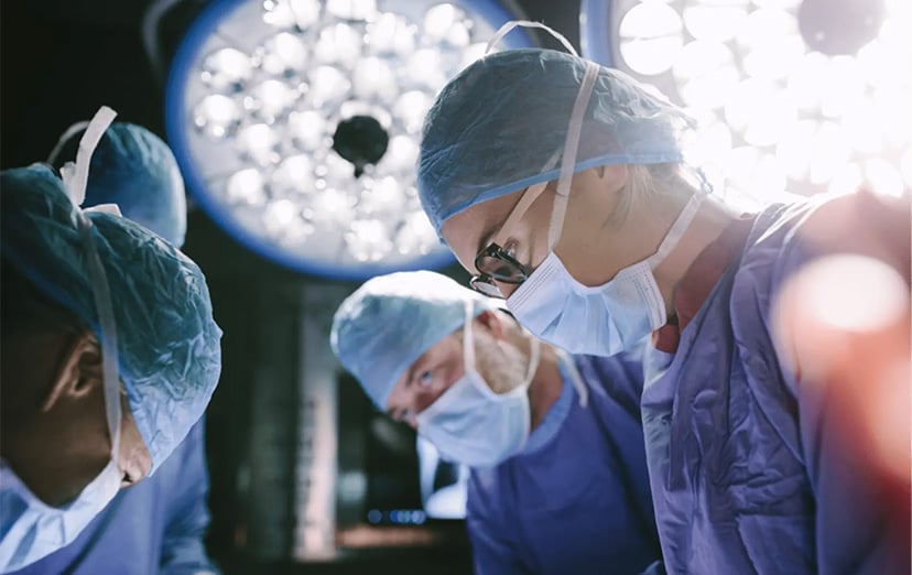 Three people in surgical gowns, caps and masks bending forward beneath operating lights