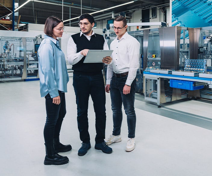Three people standing in a production hall, one holding an open laptop, machines behind them