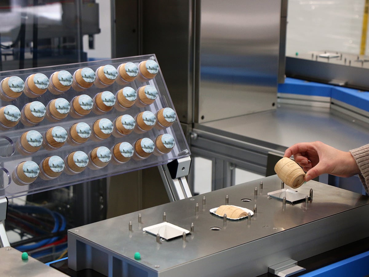 Hand placing a small wooden cylinder into a blister tray, a filled tray tilted up beside it
