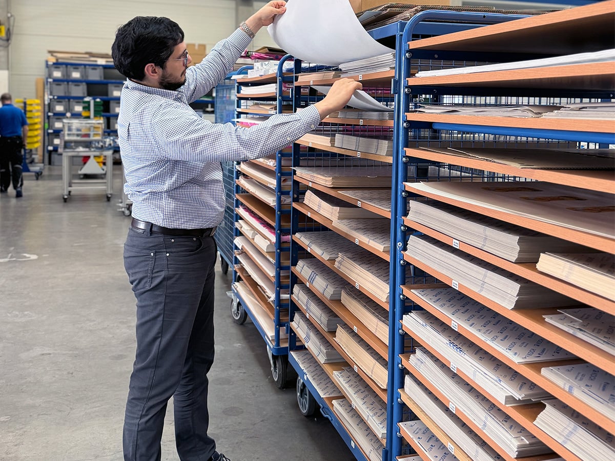 Man pulling a sheet of packaging material from a trolley with many horizontal shelves in a hall