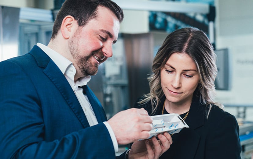 A man and a woman examining a white pack printed with the KOCH logo, machine in the background