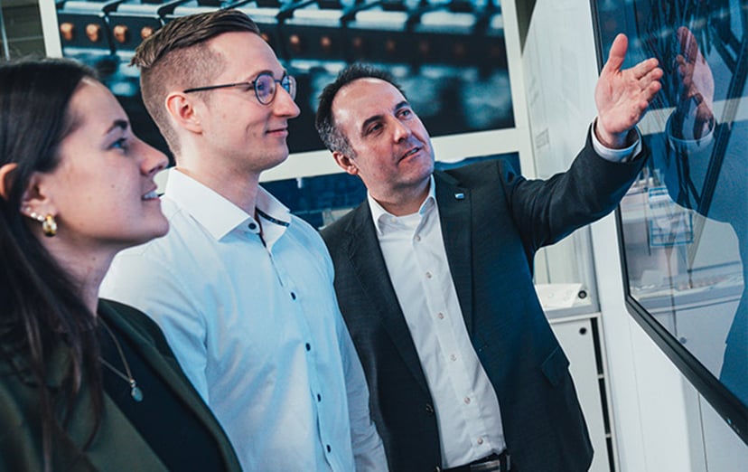 Three people in a showroom, one gesturing towards a display with machine images behind them