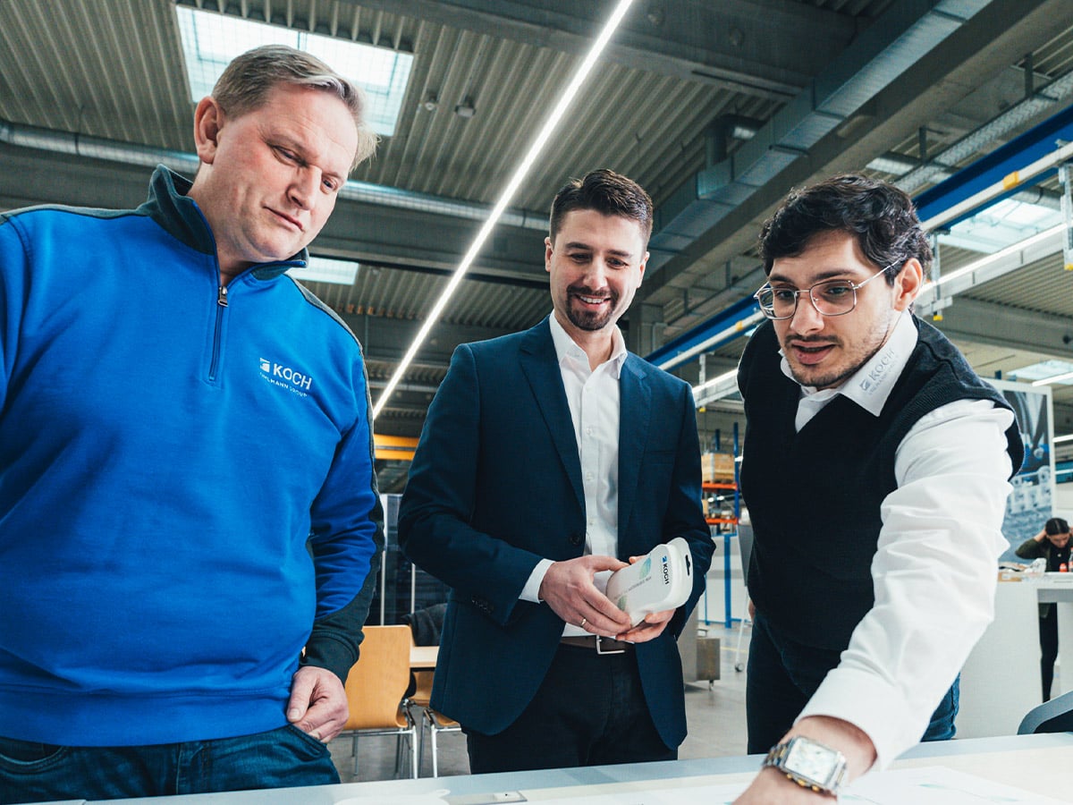 Three men at a table in a production hall, one holding a small white packaging sample