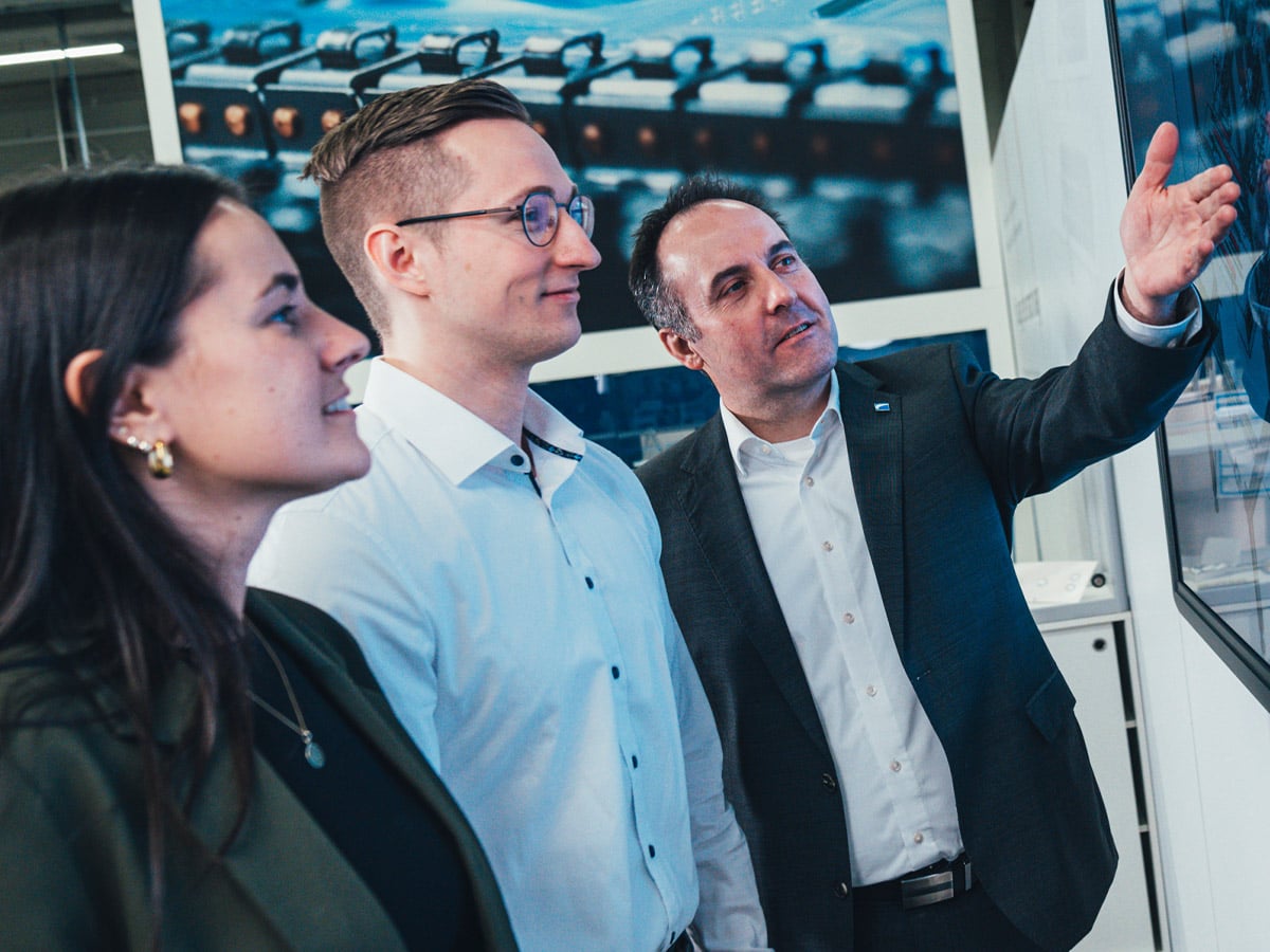 Three people in a showroom, one gesturing towards a display with machine images behind them