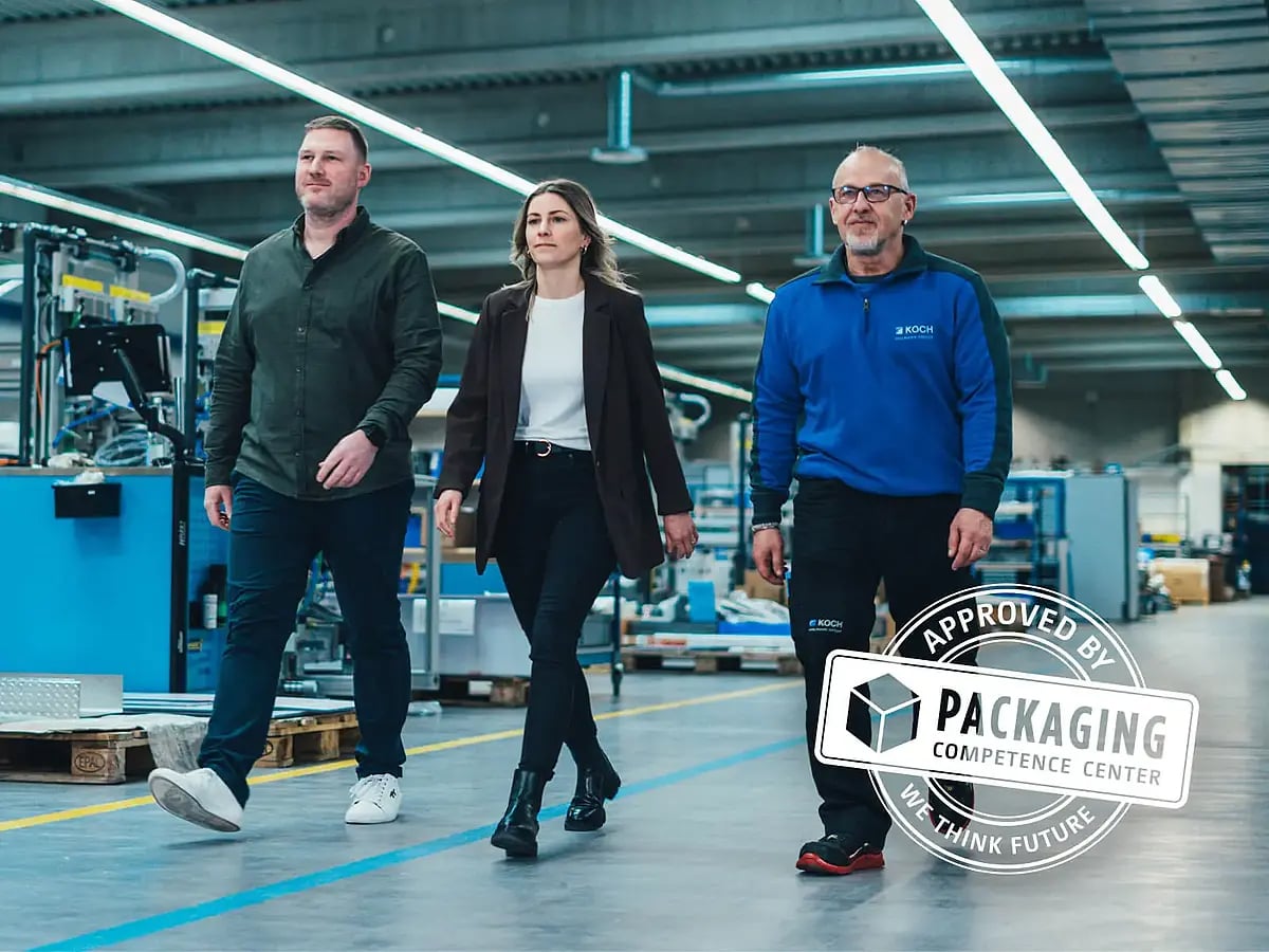Three people walking through a production hall, Packaging Competence Center approval seal at the right