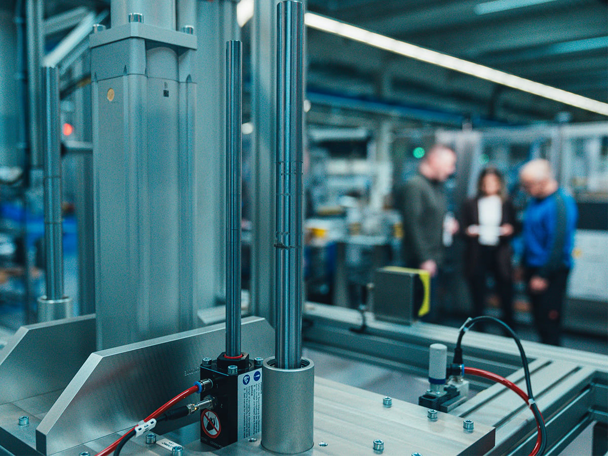 Close-up of a machine component with pneumatic cylinders, three people consulting in the blurred background