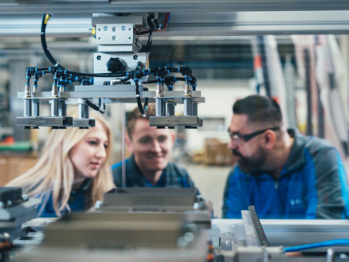 Three KOCH employees examining a pneumatic gripper head on a packaging machine in a production hall