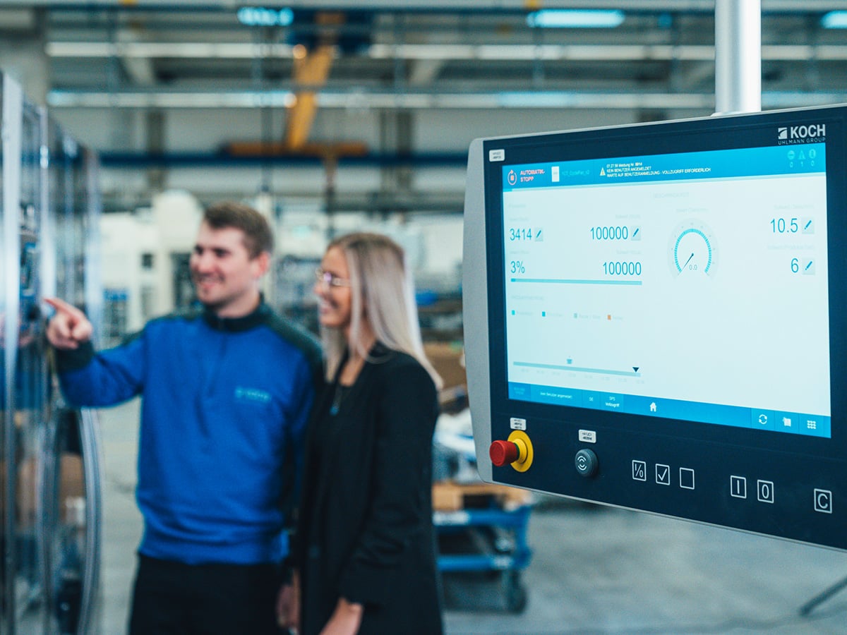 Two KOCH employees at a KOCH machine touchscreen showing process data, one pointing at the display