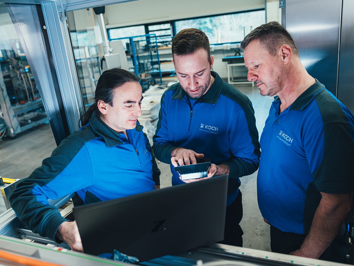 Three KOCH employees reviewing data on a laptop and tablet together at a machine station