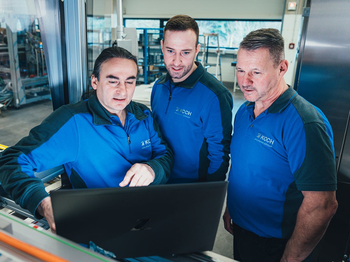 Three KOCH employees gathered around a laptop at a machine station in a production hall