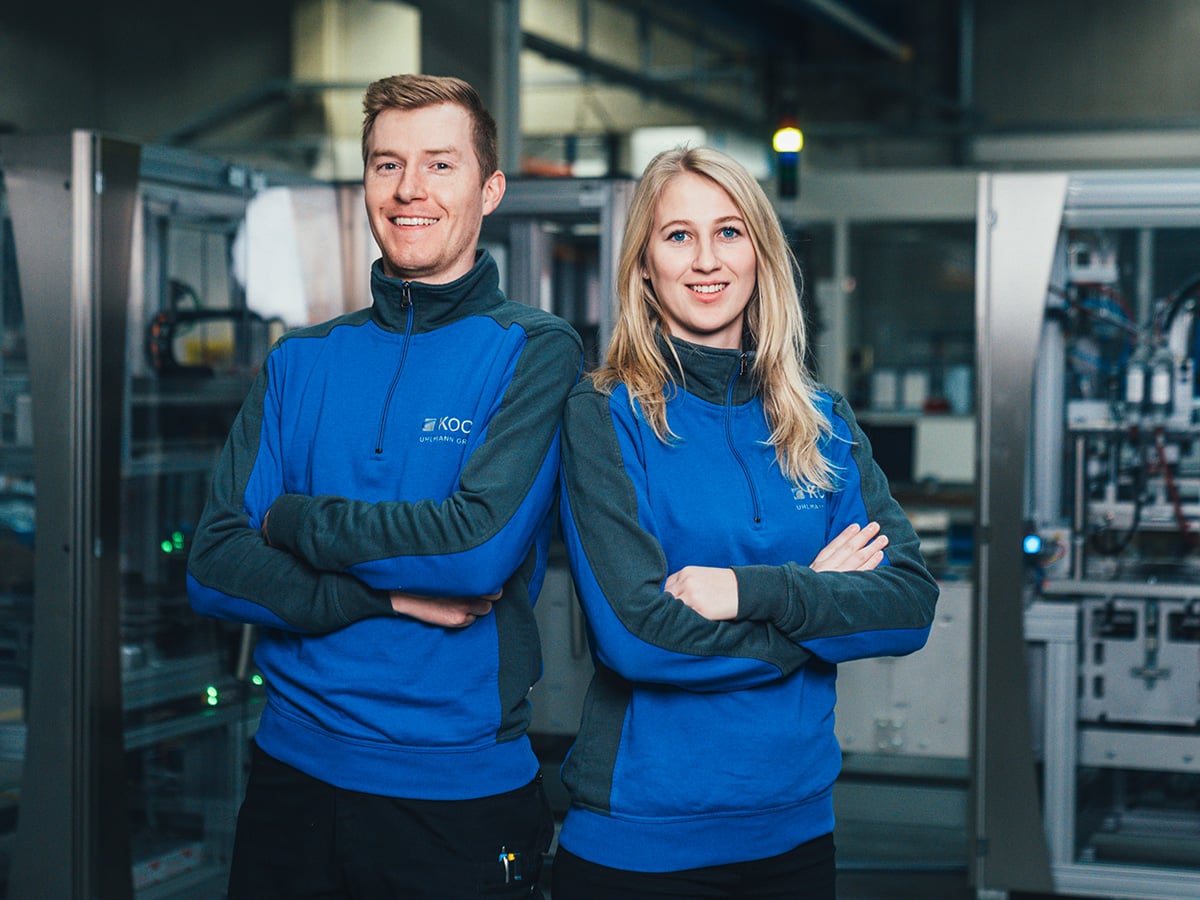 Two KOCH employees in blue work sweatshirts standing confidently with crossed arms in a production hall