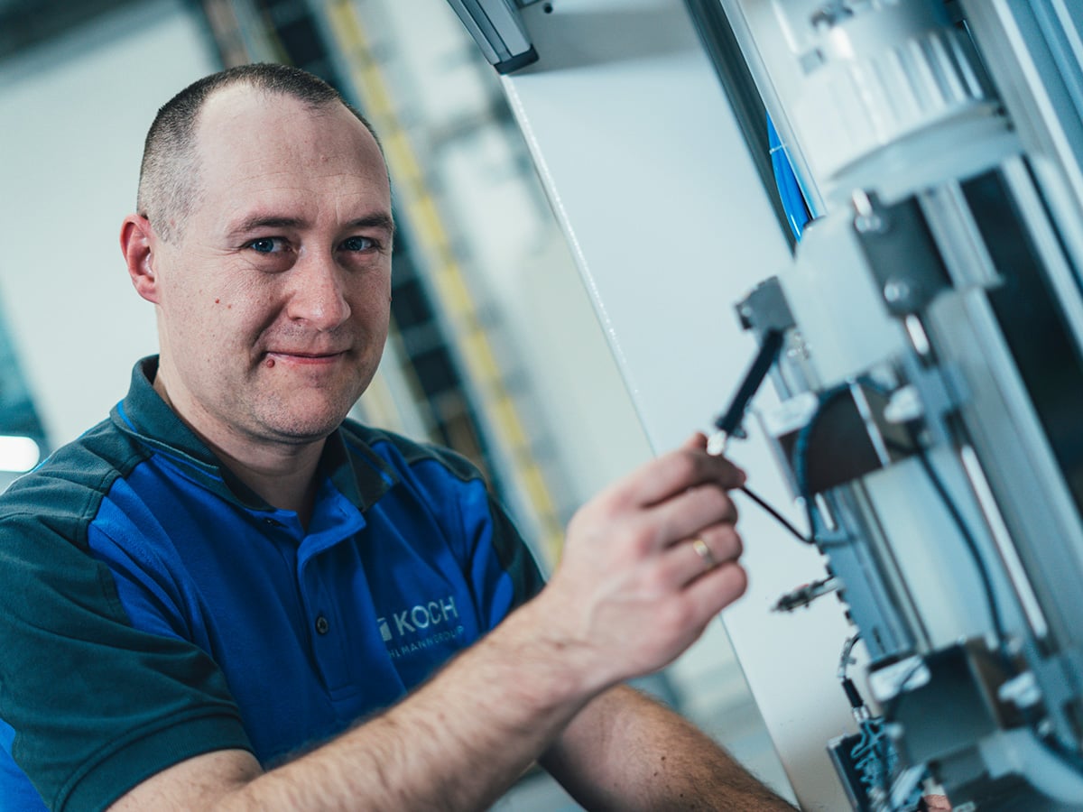 KOCH technician connecting a sensor cable to a machine component, smiling at the camera