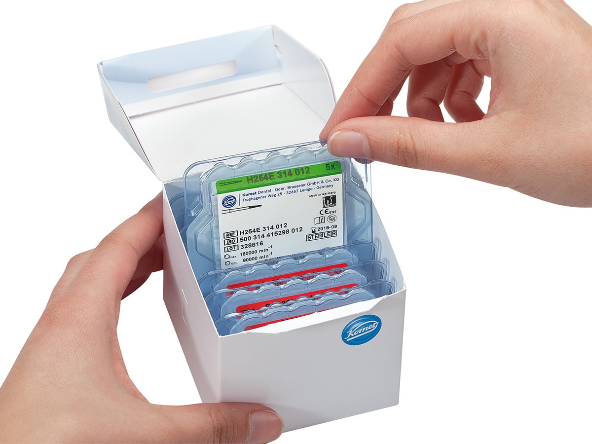 Hands opening a white Komet carton holding several upright transparent blisters with printed labels