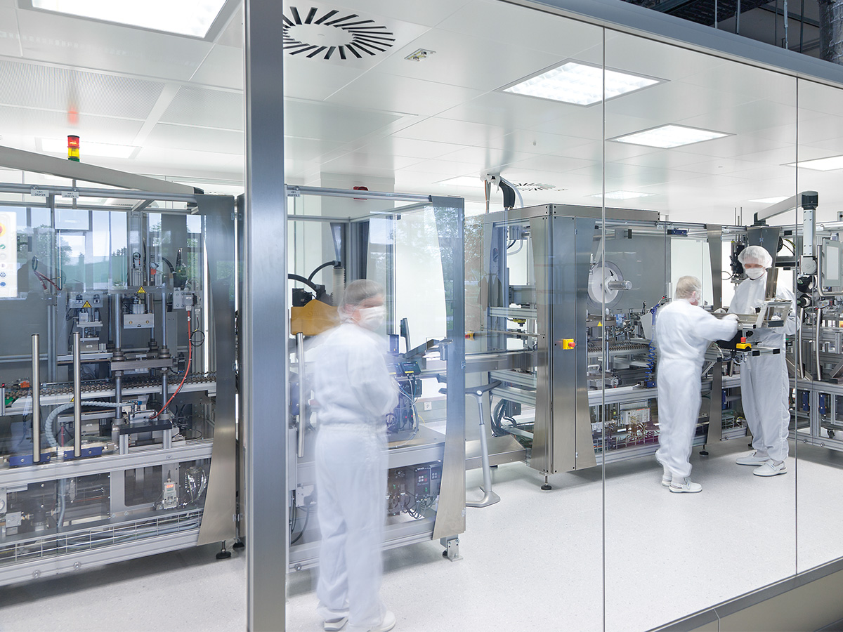 Three people in cleanroom garments working at an automated packaging line behind glass walls