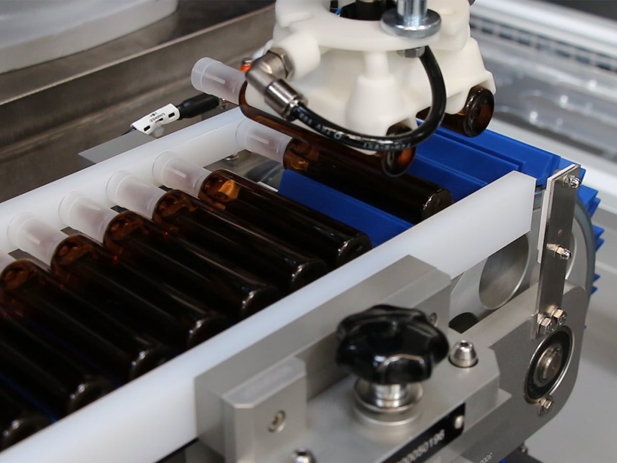 Gripper head lifting two brown glass ampoules from a row of white guide channels