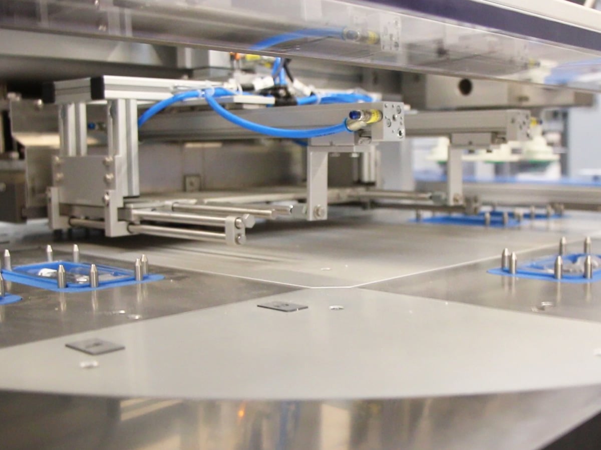 Rotating table with blue holders carrying flat blisters beneath a gantry with suction grippers