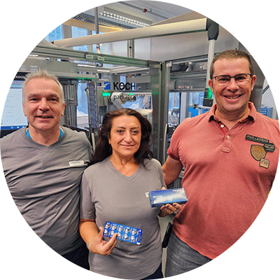 Thomas Meier of Renata with two colleagues holding button cell blister packs in front of a KOCH machine