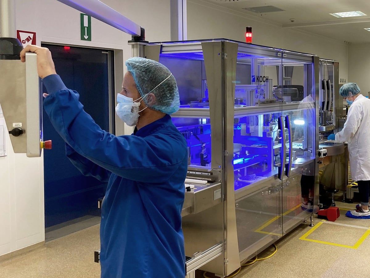 Two people in cleanroom garments operating a KOCH KDT packaging machine with blue UV lighting