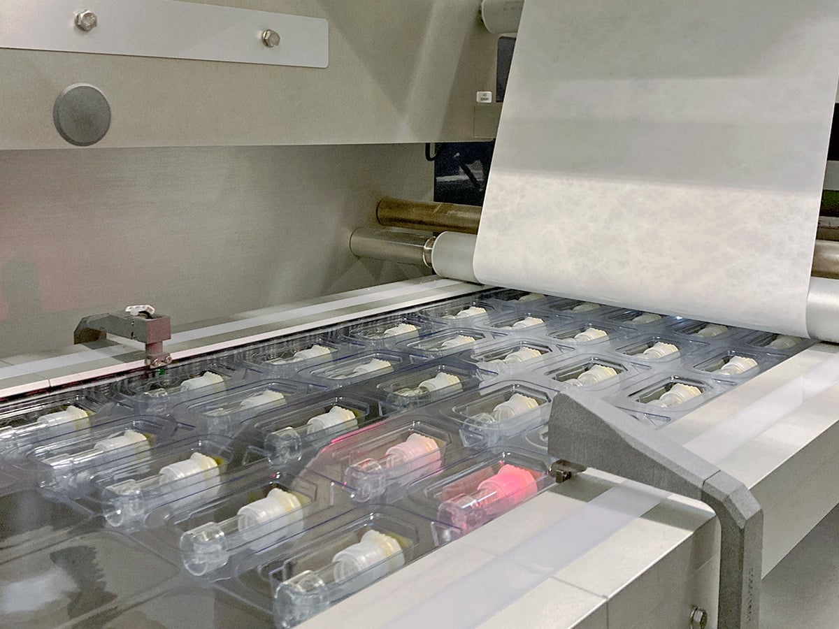 Rows of vials in blister trays entering the sealing station as a white lidding web closes over them