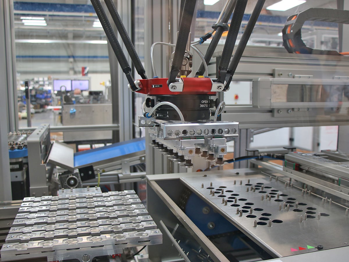 Delta robot arm lowering onto a plate with rows of button cell cavities between the two blister machines