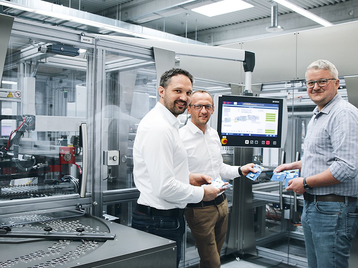Three people holding VARTA button cell blister packs in front of a KOCH packaging machine