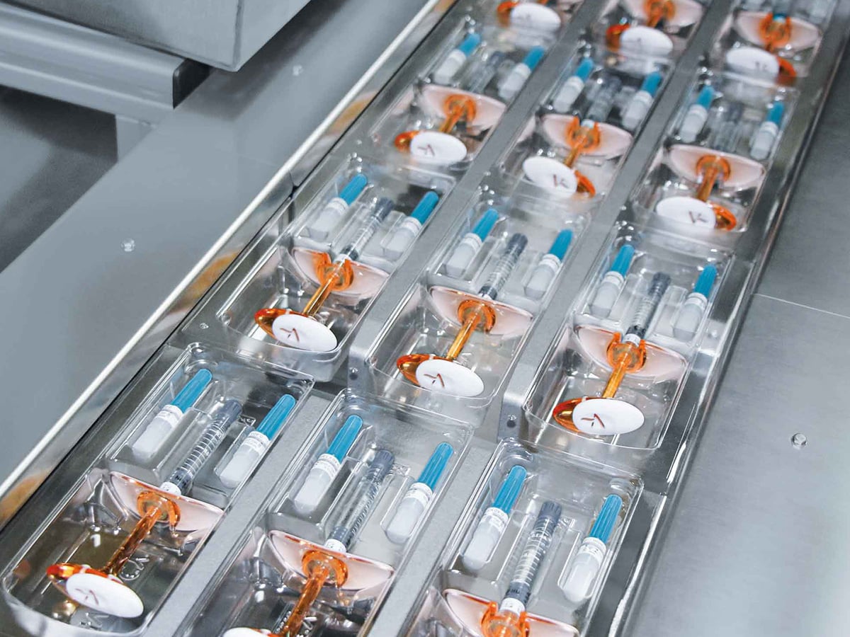 Two rows of prefilled syringes with orange caps in transparent blister trays on a conveyor