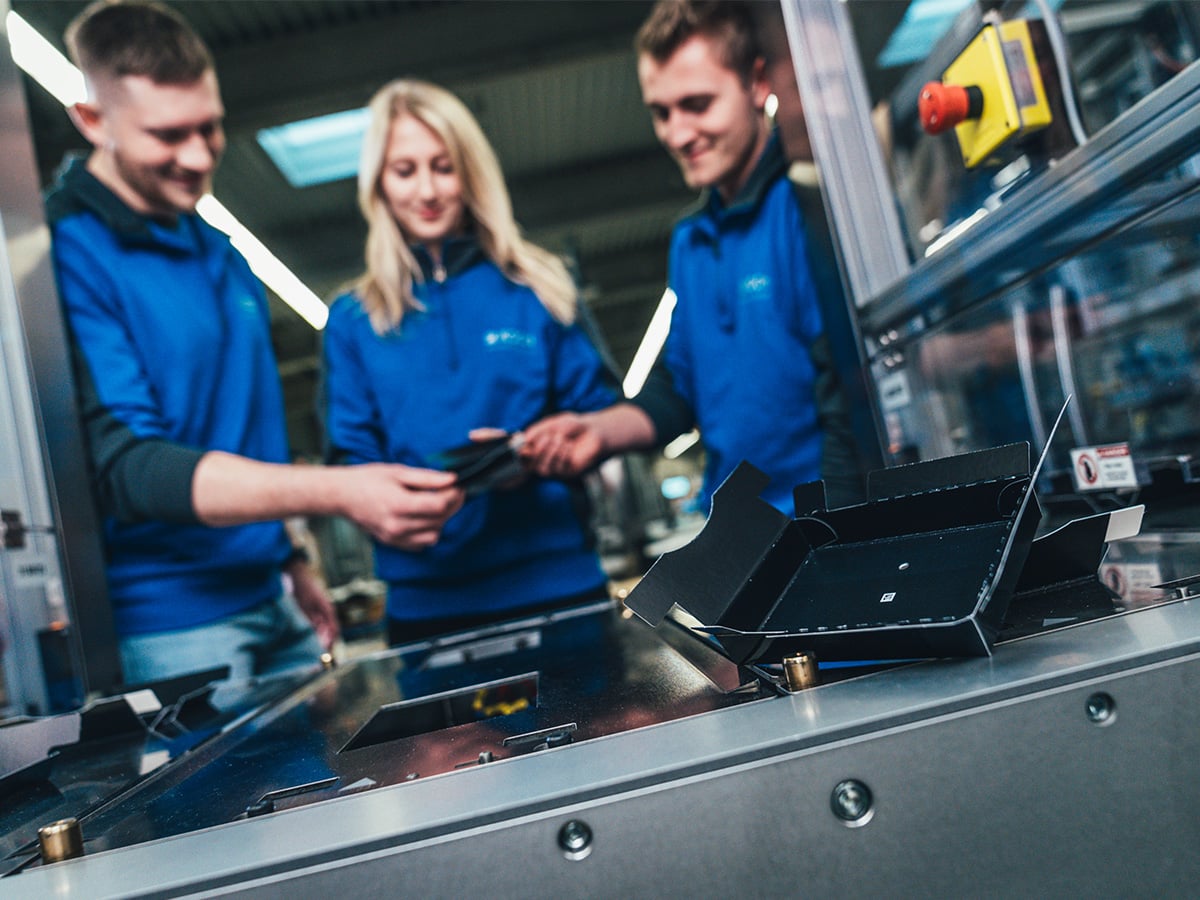 Three KOCH Pac-Systeme employees in blue workwear operating a packaging machine with a tablet, illustrating innovation in practice