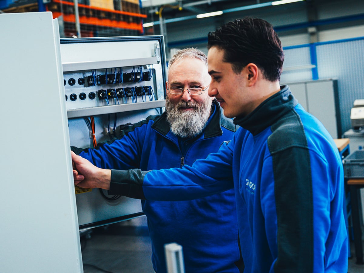 Two KOCH Pac-Systeme employees in blue workwear working together on an electrical control cabinet in a production hall