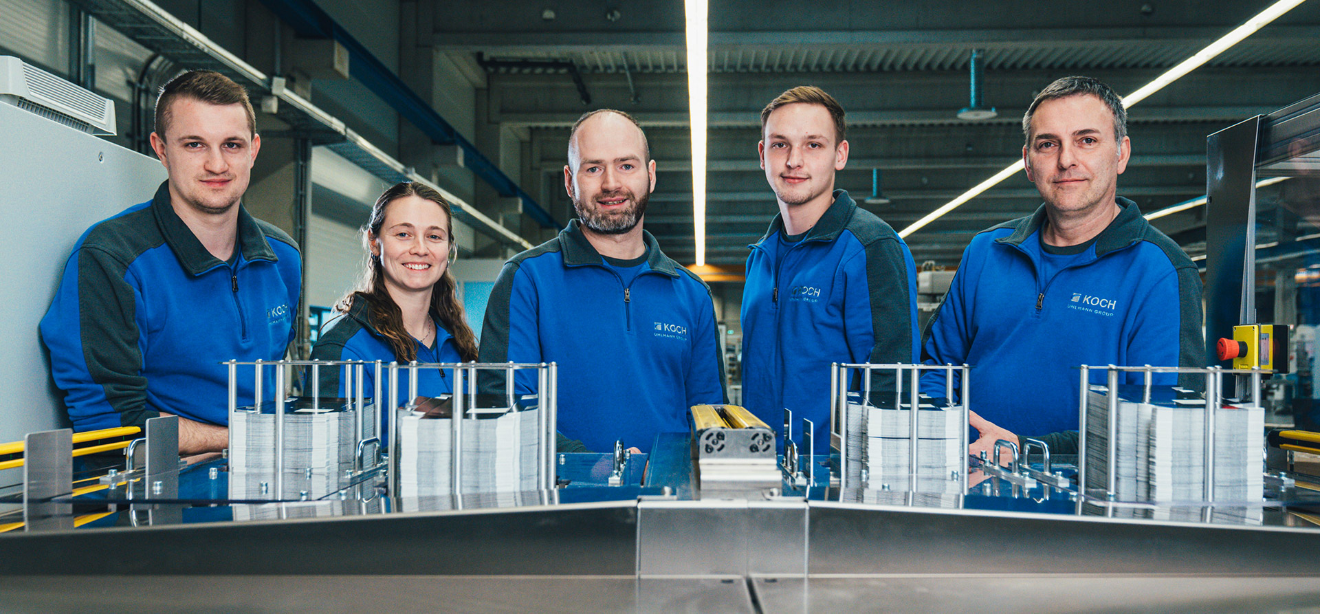 Five KOCH Pac-Systeme employees in blue workwear posing together at a packaging machine in a production hall