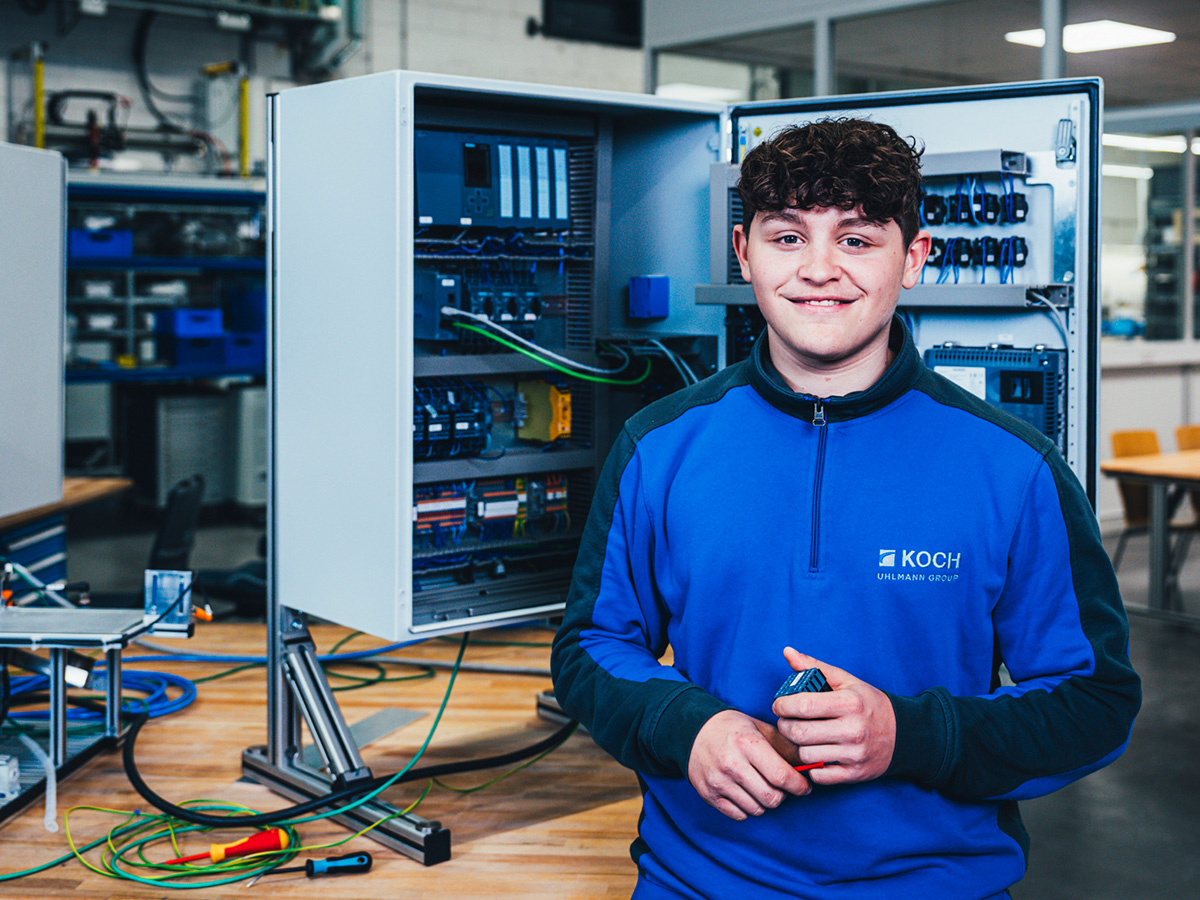 Young KOCH apprentice in blue workwear standing in front of an open electrical control cabinet in a training workshop, representing the Electronics Technician for Automation Technology apprenticeship