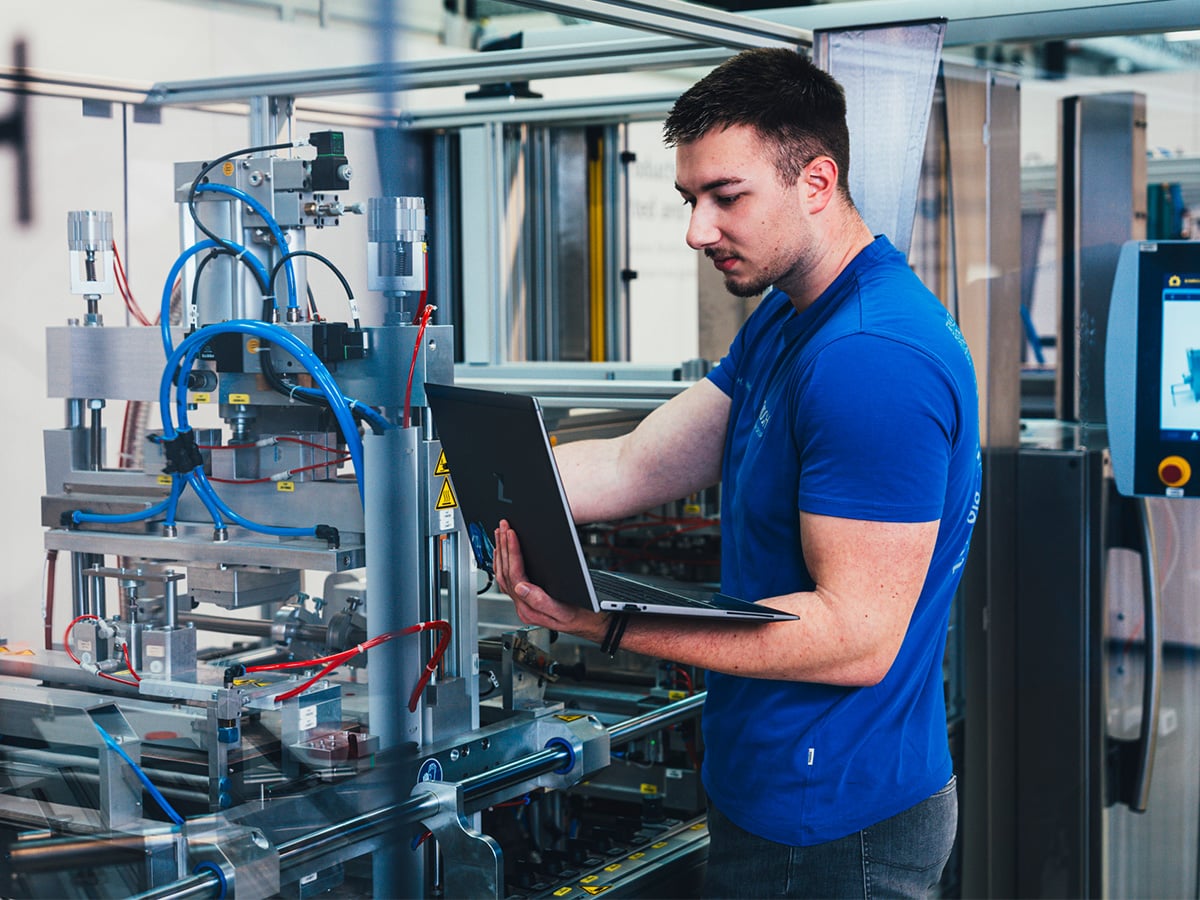 Dual study student in blue KOCH workwear using a laptop at a packaging machine, representing the Bachelor of Engineering in Mechanical Engineering programme