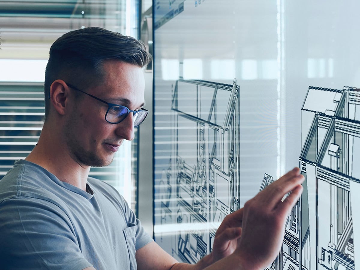 Young man with glasses examining a technical engineering drawing on a glass panel, representing the Bachelor of Engineering in Industrial Engineering programme at KOCH Pac-Systeme