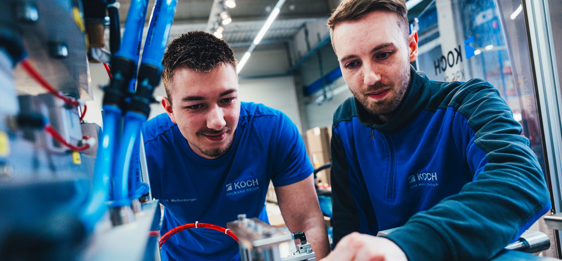 Two KOCH Pac-Systeme interns in blue workwear working together on a packaging machine in a production environment
