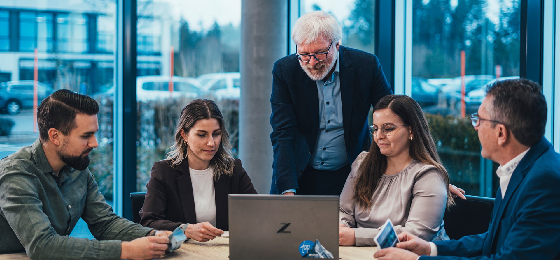 Fünf KOCH Pac-Systeme Mitarbeiter in einem gemeinsamen Meeting an einem Tisch mit Laptop in einer modernen Büroumgebung mit großen Fenstern