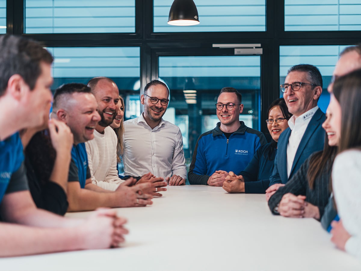 Diverse group of KOCH Pac-Systeme employees laughing together at a large table in a modern meeting room