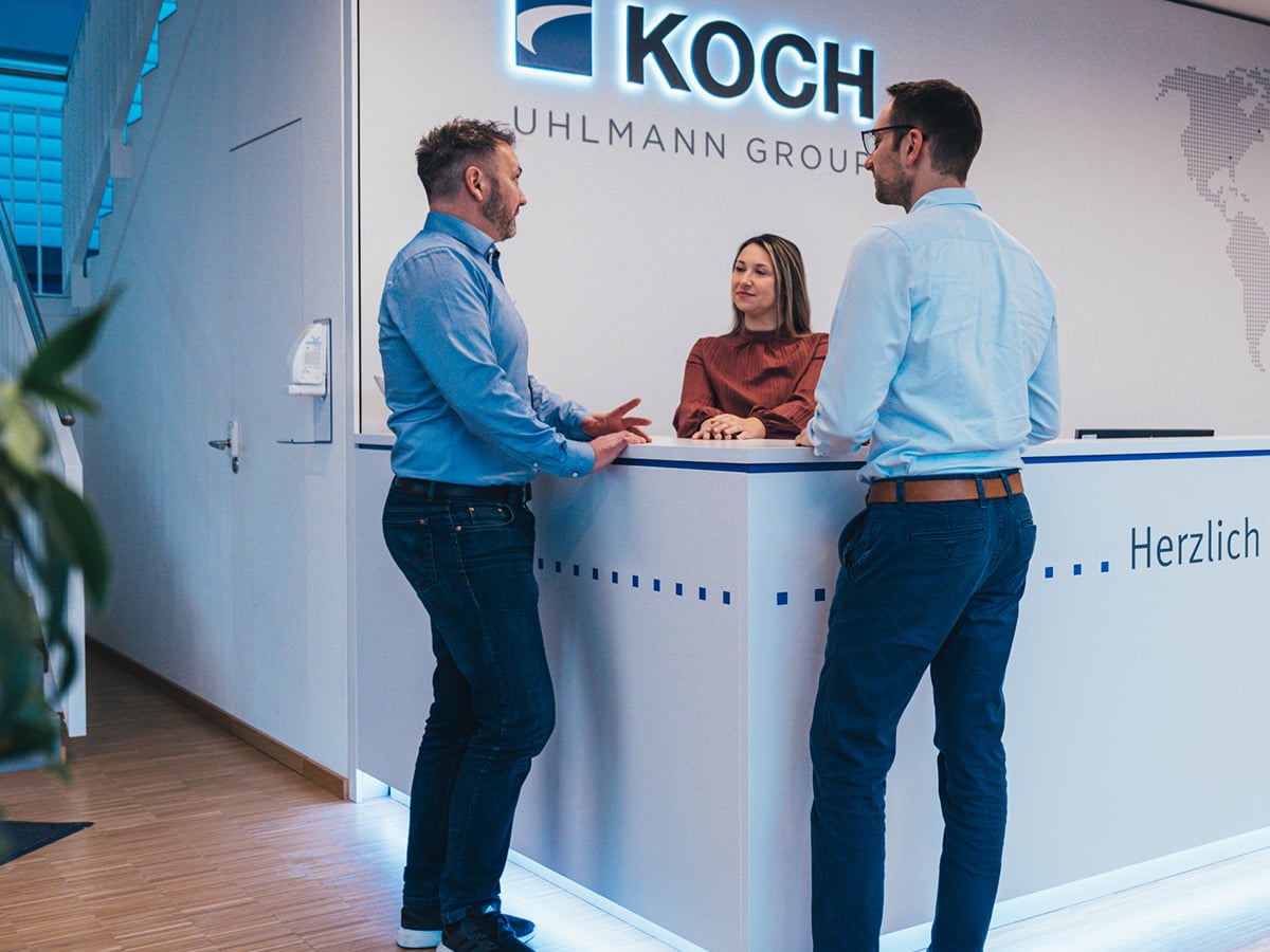 Three KOCH Pac-Systeme employees in conversation at the company reception, with the illuminated KOCH Uhlmann Group logo visible in the background